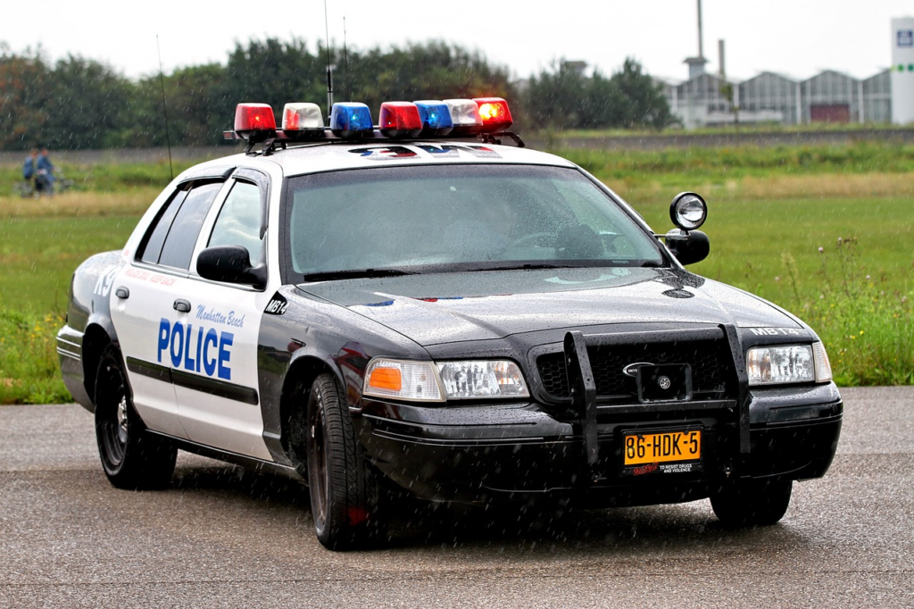 An image capturing the aftermath of an incident in Anacortes, Washington: police vehicles surround a mangled car on a rain-soaked street, flashing lights reflecting off shattered glass and twisted metal